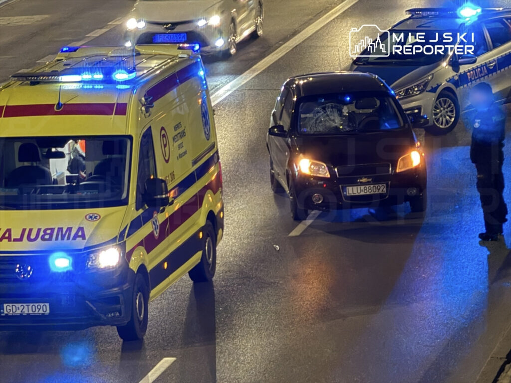 Karetka pogotowia z włączonymi sygnałami świetlnymi zatrzymana obok uszkodzonego samochodu osobowego na ruchliwej ulicy.