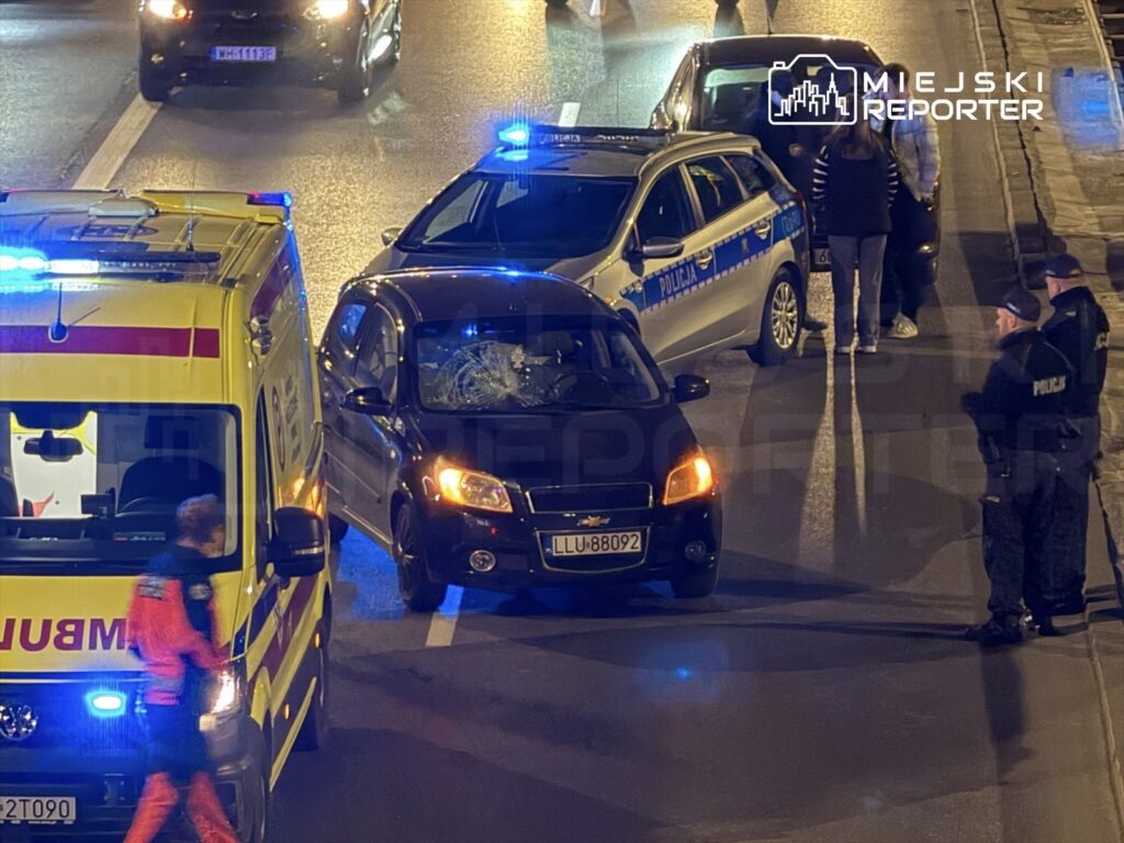 Na jezdni stoi czarny samochód z uszkodzoną szybą, obok niego policyjny radiowóz oraz karetka pogotowia. W tle widoczni funkcjonariusze rozmawiają z osobami.