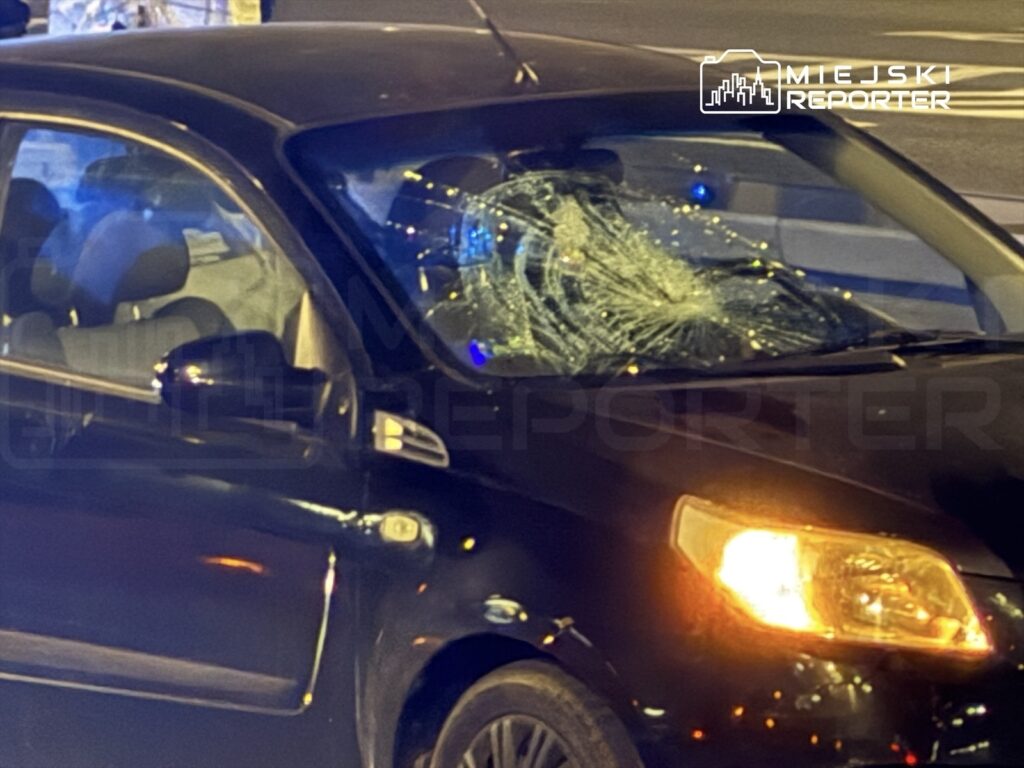 Czarny samochód osobowy z rozbitą szybą przednią stoi na ulicy w nocy, otoczony przez światła policyjne.