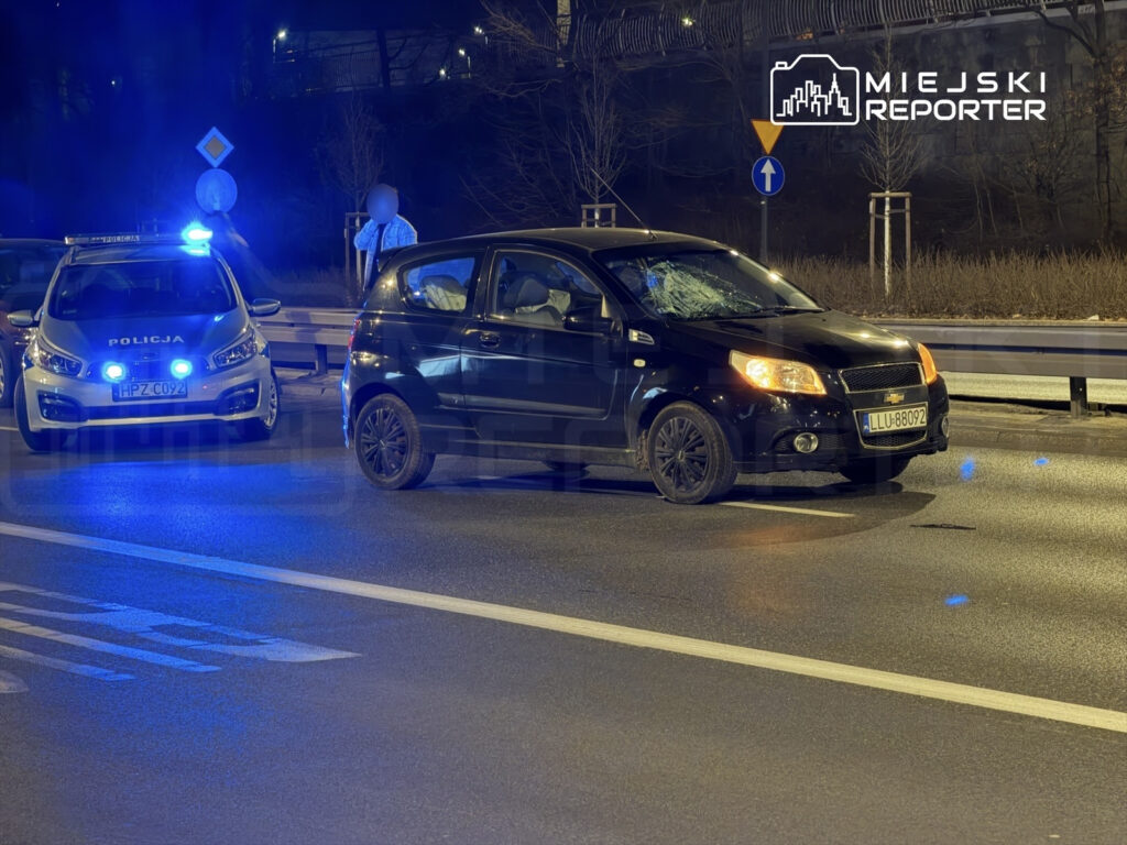 Czarny samochód osobowy z uszkodzoną szybą stoi na drodze, obok policyjny radiowóz z włączonymi sygnałami świetlnymi.