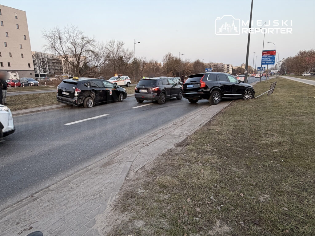 Trzy uszkodzone pojazdy na jezdni, w tym taksówka z wgniecioną boczną częścią, obok zaparkowany czarny SUV.