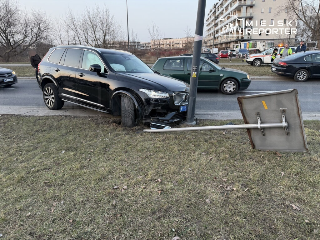 Czarny SUV wjeżdża na trawnik, uszkadzając słup z oznakowaniem drogowym. W tle widoczne inne pojazdy i budynki.
