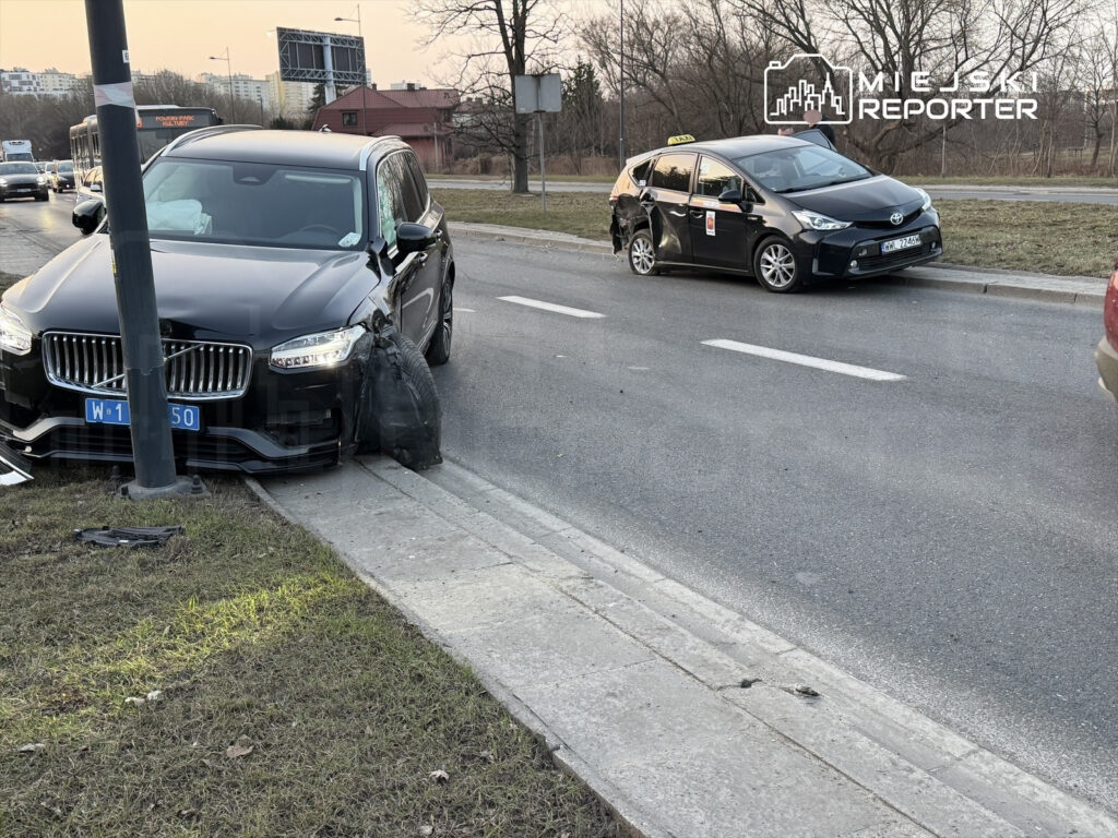 Czarny SUV uderzył w słup, stojąc na chodniku, obok taksówka z widocznymi uszkodzeniami.
