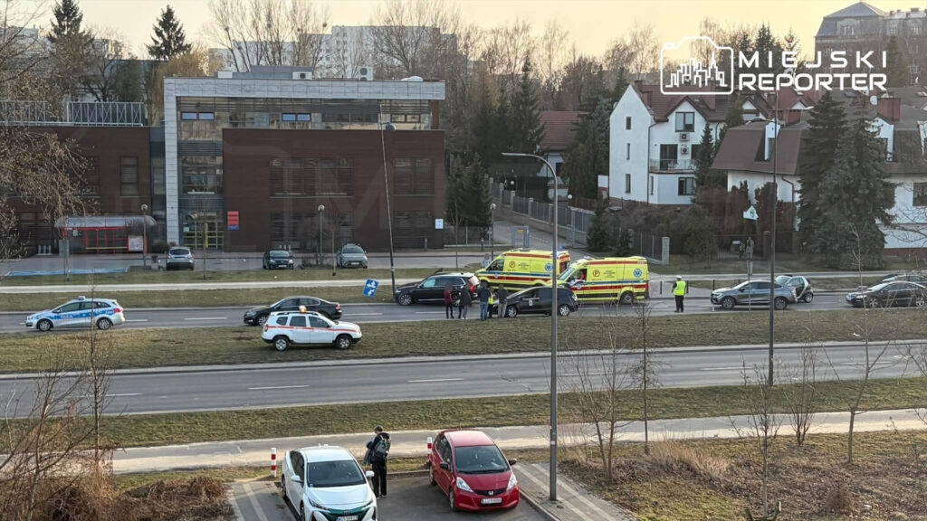 Na jezdni zatrzymane pojazdy, w tym ambulansy, oraz grupa osób rozmawiająca obok. W tle nowoczesny budynek i domy.