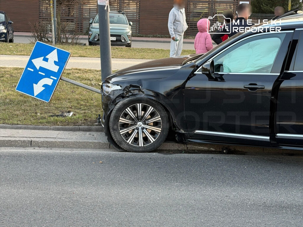 Czarny samochód osobowy z uszkodzoną przednią częścią stoi obok przewróconego znaku drogowym na chodniku.