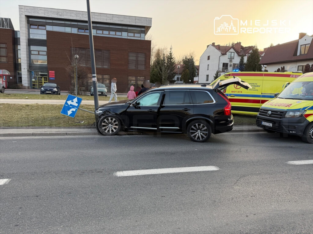 Czarny SUV zaparkowany obok przewróconego znaku drogowym, obok stoi karetka pogotowia, w tle przechodnie.