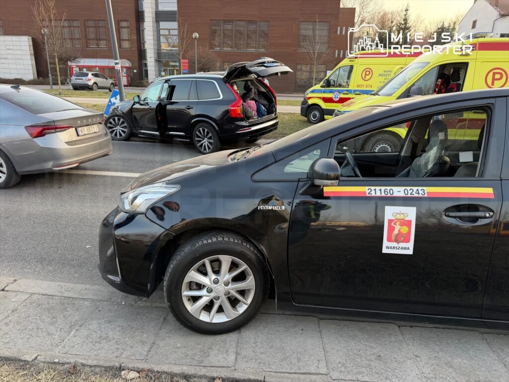 Czarny samochód osobowy z logo Warszawy stoi na poboczu, obok otwartego SUV-a, w tle widoczne karetki pogotowia.