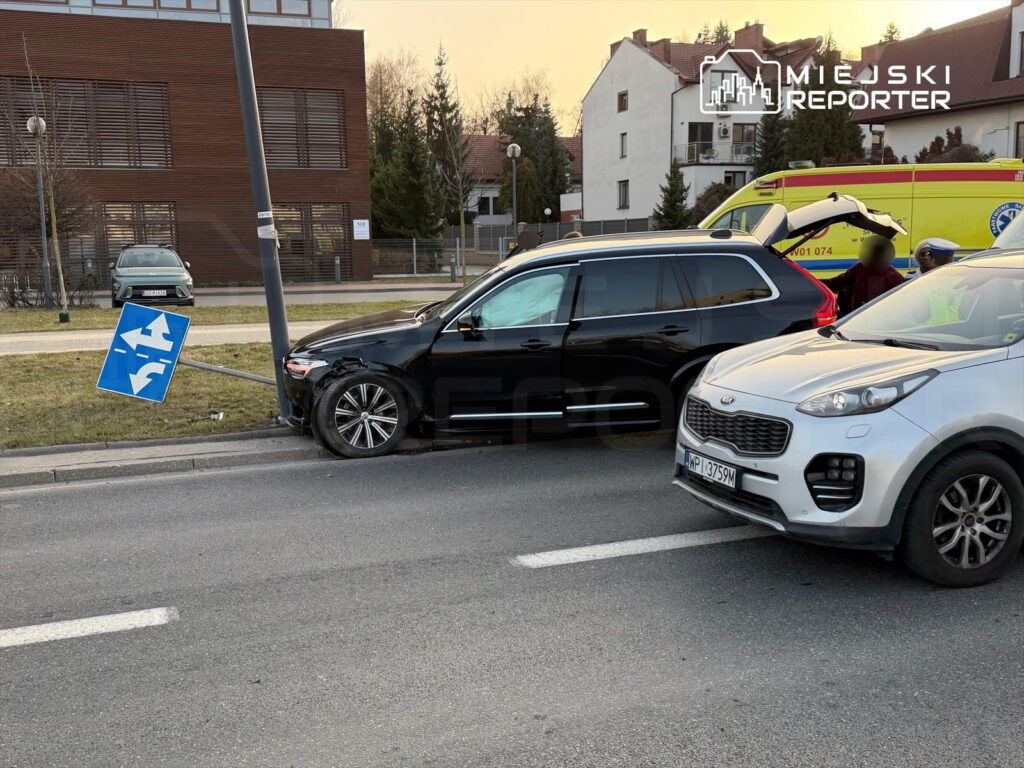 Czarny SUV uderzył w znak drogowy, który opiera się na słupie, obok białego samochodu w miejskim otoczeniu.