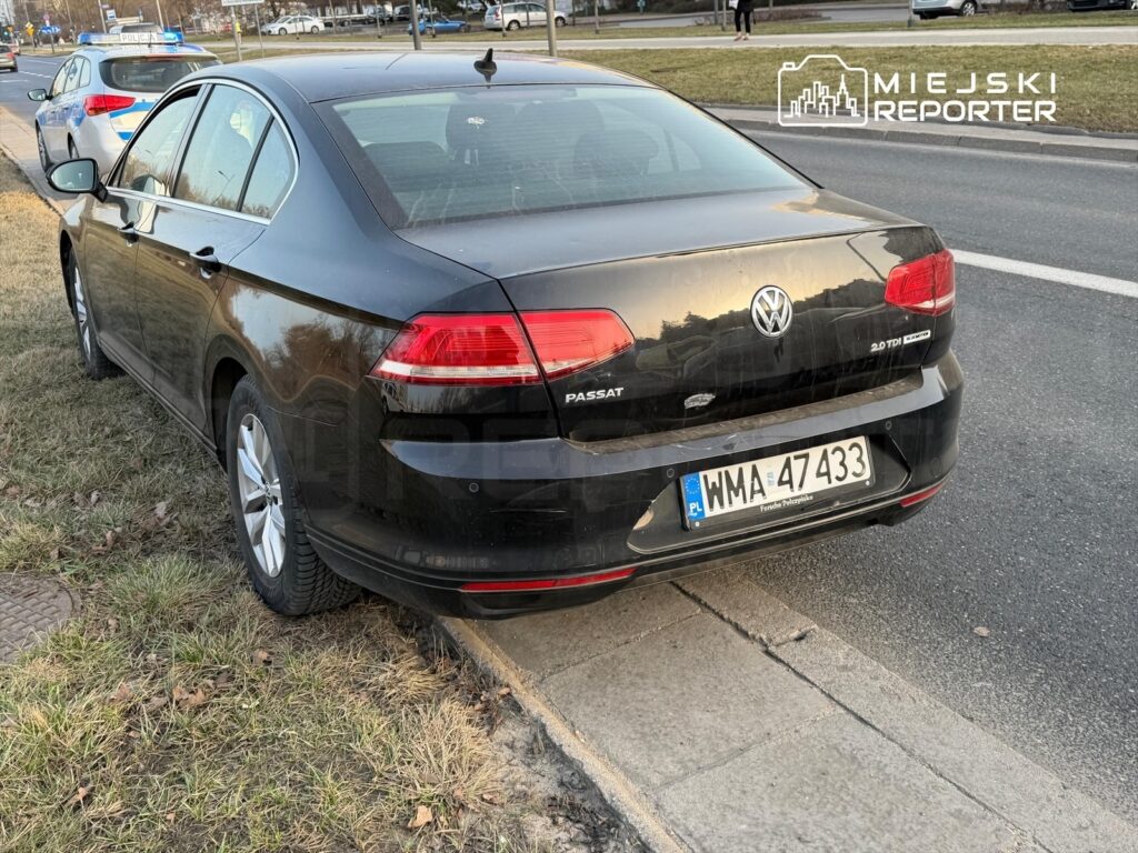 Czarny Volkswagen Passat zaparkowany na chodniku obok policyjnego radiowozu na miejskiej ulicy.