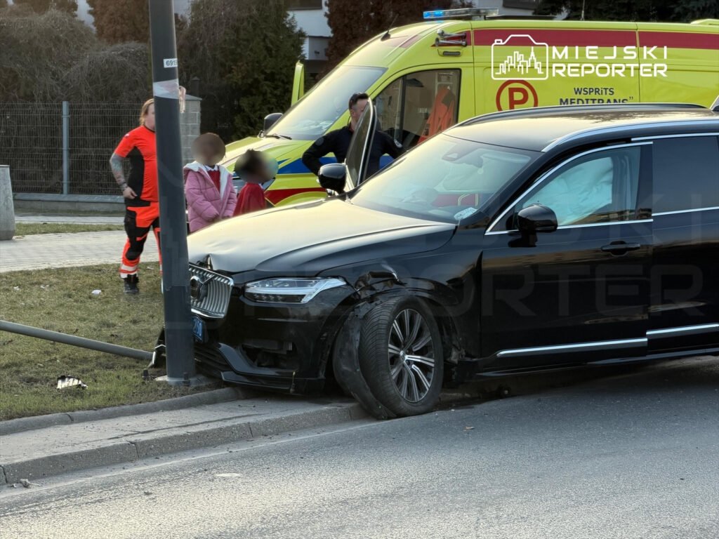 Czarny samochód osobowy uderzył w słup, a obok stoi ratownik medyczny z dziećmi w pobliżu. W tle widoczna karetka.
