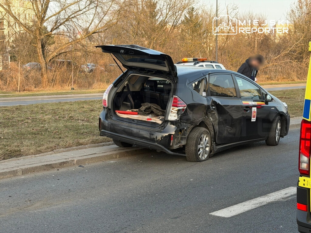 Czarny samochód osobowy z uszkodzoną tylną częścią stoi na poboczu drogi, obok pracownik sprawdzający sytuację.