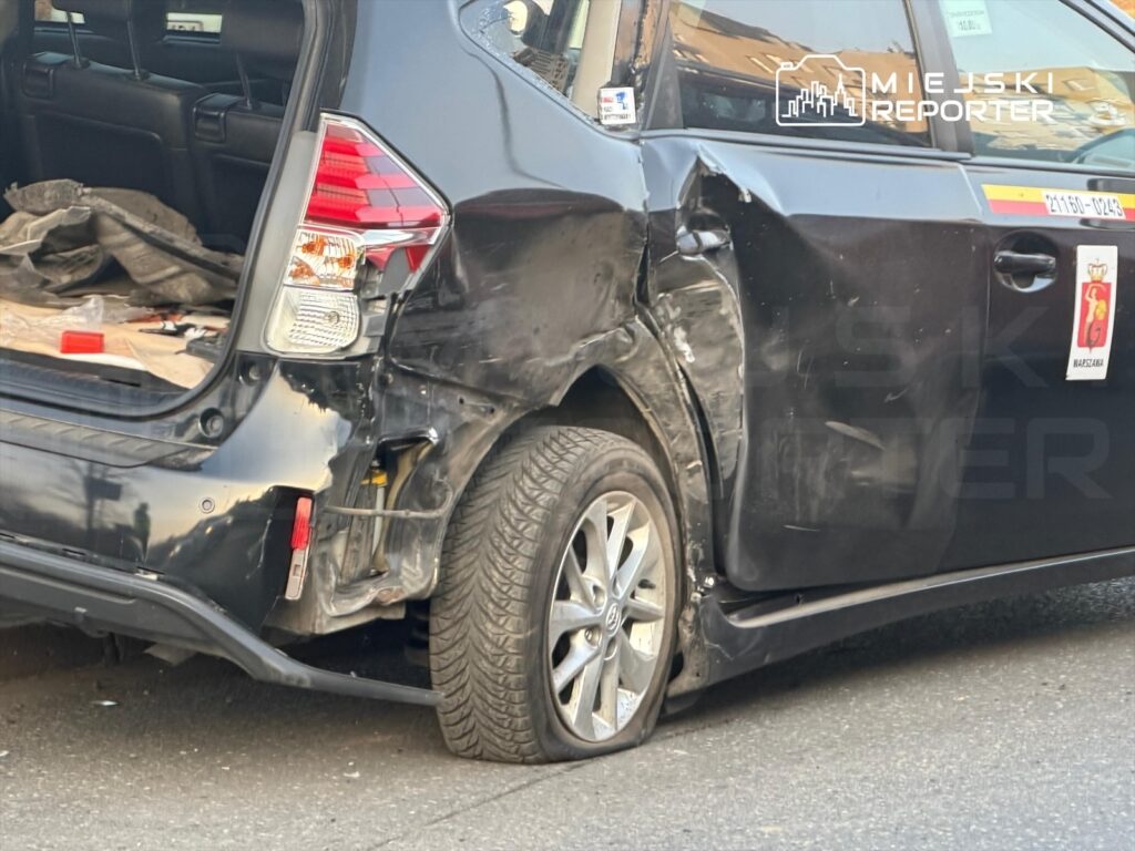 Czarny samochód osobowy z uszkodzeniami tyłu, zaparkowany na ulicy, z widoczną przebitą oponą.