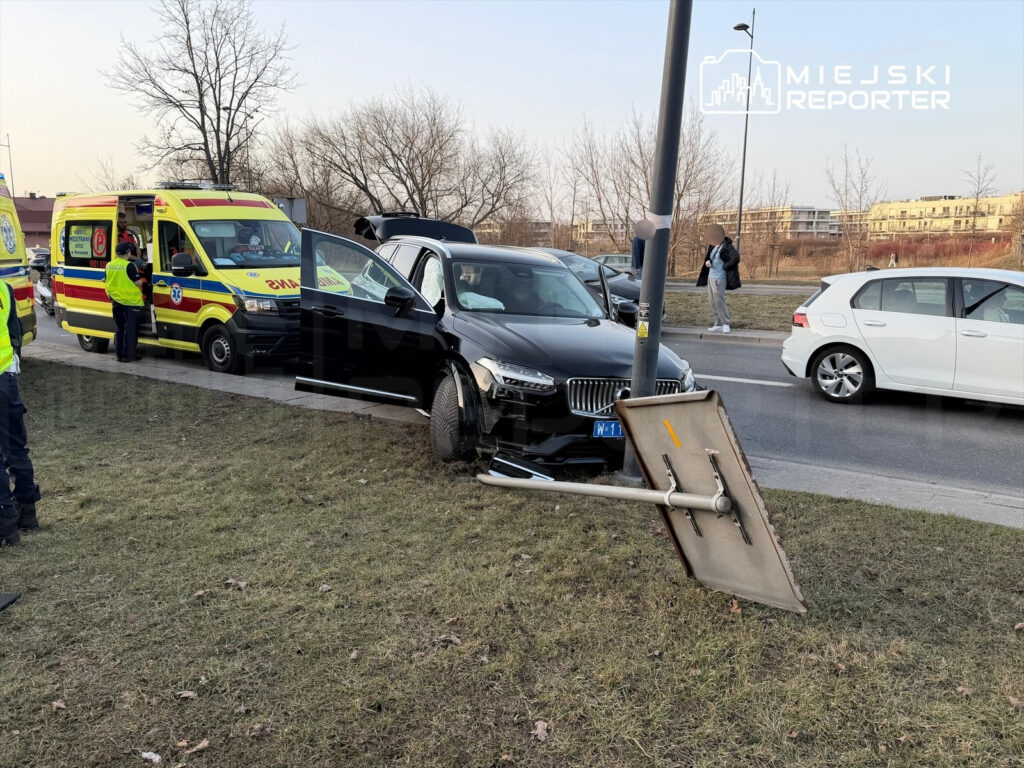 Czarny samochód osobowy uderzył w znak drogowy, obok stoi karetka i ratownicy medyczni rozmawiają z kierowcą.