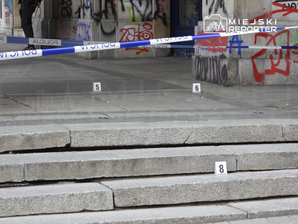 Policjant stojący przy schodach z oznaczeniami miejsc zdarzenia, otoczenie pokryte graffiti.