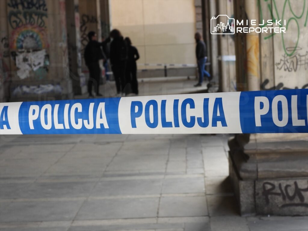 Policja wyznaczyła strefę bezpieczeństwa taśmą, a w tle widoczni przechodnie w miejskim otoczeniu.