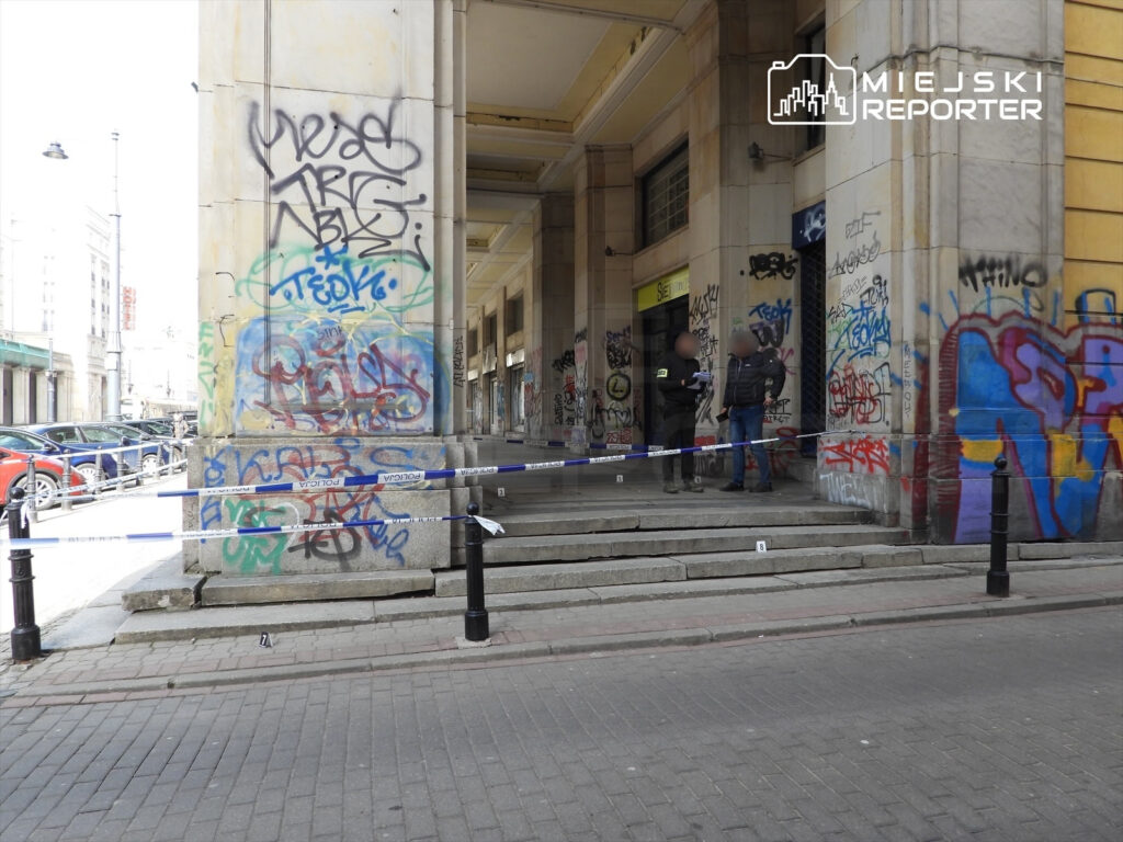 Dwaj policjanci rozmawiają przy zablokowanych schodach budynku z graffiti, otoczeni taśmą policyjną.