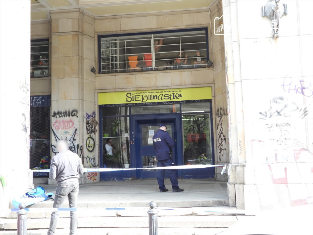 Mężczyzna w szarym swetrze stoi przed wejściem do budynku z widocznymi graffiti, obok policjant przeszukuje okolicę.