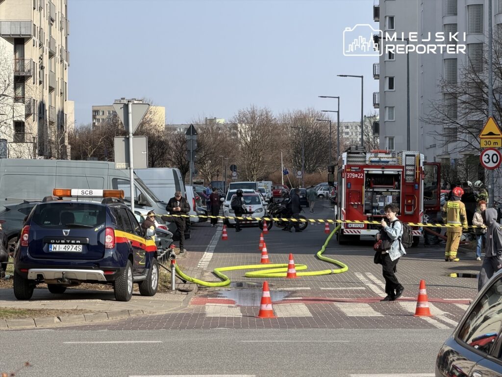 Na ulicy, w otoczeniu bloków mieszkalnych, zespół strażaków pracuje przy wozie strażackim, a obok ustawione są służby ratunkowe.