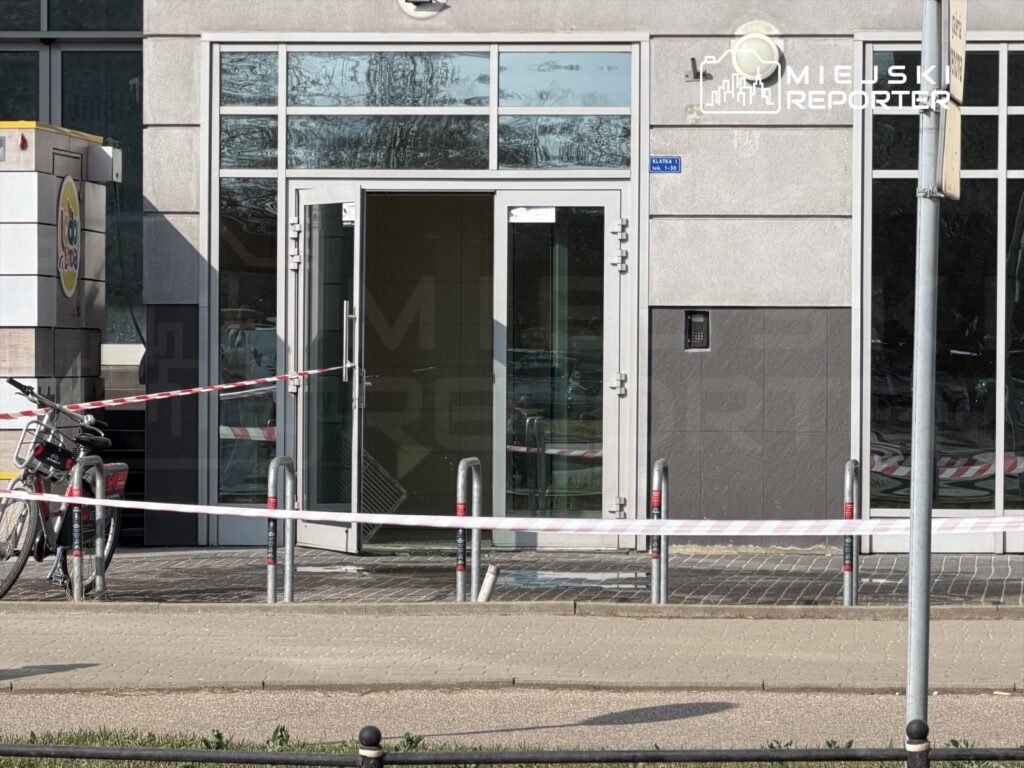Wejście do budynku z przeszkloną fasadą zabezpieczone taśmą, obok rower na stojaku.