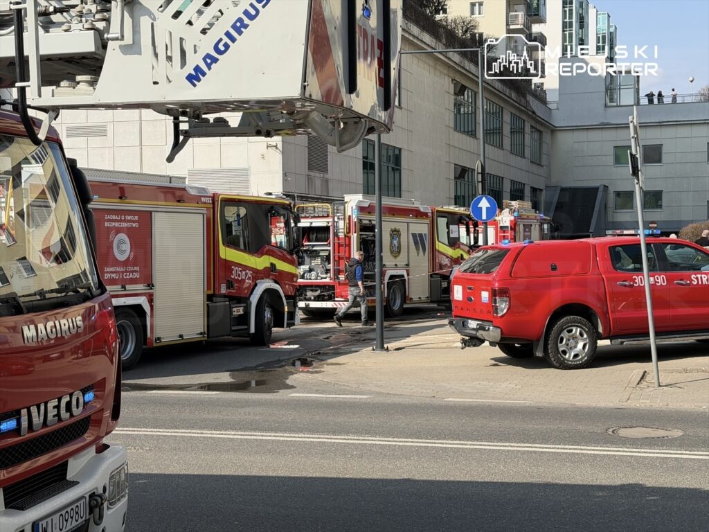 Strażacy przy wozach strażackich w pobliżu budynku, w tle widoczna podnośnik i dodatkowe pojazdy ratunkowe.