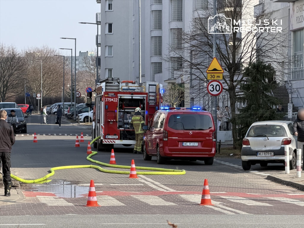 Strażacy przy wozie strażackim z wężem pożarniczym, w tle samochody oraz przechodnie na ulicy.