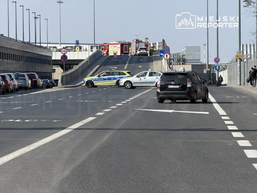 Policyjny radiowóz blokujący ruch na drodze prowadzącej do parkingu, obok zaparkowane samochody i w tle wjeżdżające pojazdy.