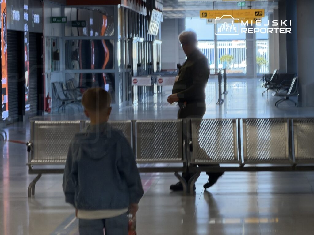 Chłopiec w dżinsowej kurtce trzyma butelkę wody, idzie w kierunku mężczyzny w mundurze w nowoczesnym obiekcie.