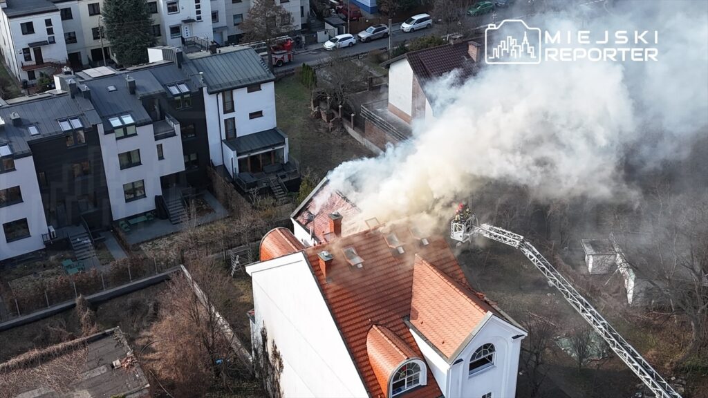 Dym wydobywający się z dachu budynku mieszkalnego, podczas akcji gaśniczej prowadzonej przez strażaków na podnośniku.