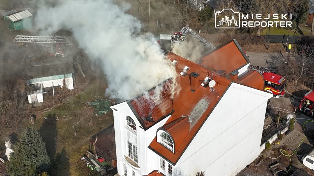 Dym wydobywający się z dachu białego budynku mieszkalnego, w tle widać strażacki wóz i drabinę.