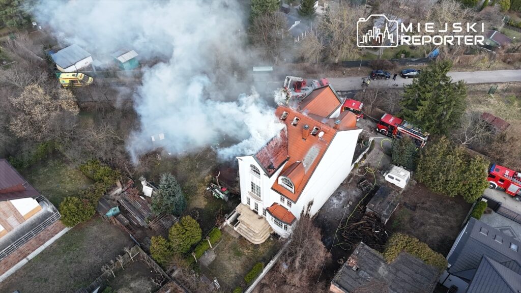Ogromne kłęby dymu unoszą się z dachu nowoczesnego budynku mieszkalnego, wokół którego znajdują się wozy strażackie.