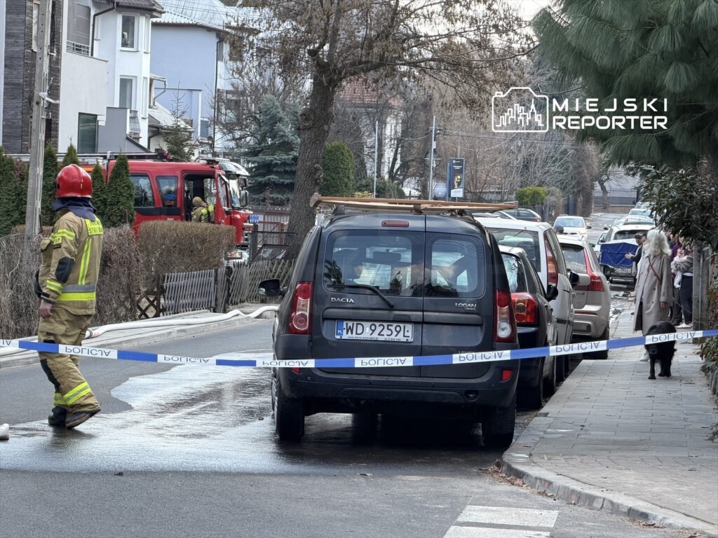 Strażak w hełmie kontroluje sytuację na miejscu zdarzenia, obok redakcyjny wóz strażacki, a ulica jest zabezpieczona taśmą.