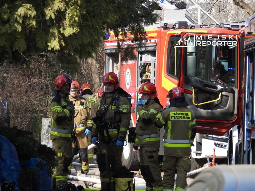 Grupa strażaków w mundurach ochronnych rozmawia obok wozu strażackiego, w tle widoczne są sprzęty ratunkowe.