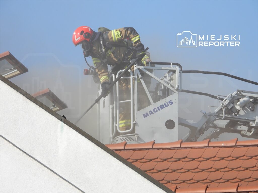 Strażak w hełmie i maskach ochronnych pracuje na dźwigu strażackim, gasząc pożar na dachu budynku.