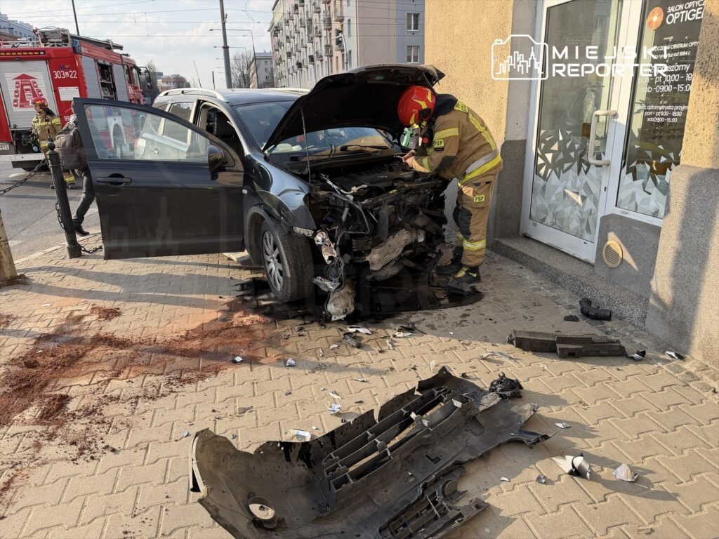 Strażak w kasku i mundurze naprawia uszkodzony samochód osobowy zaparkowany przy budynku, obok leży uszkodzona część pojazdu.