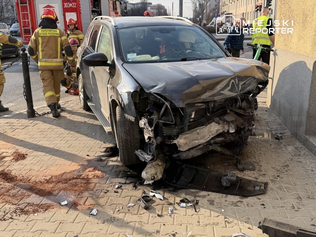 Czarny samochód osobowy z uszkodzonym przodem stoi na chodniku, obok pracują strażacy w mundurach.