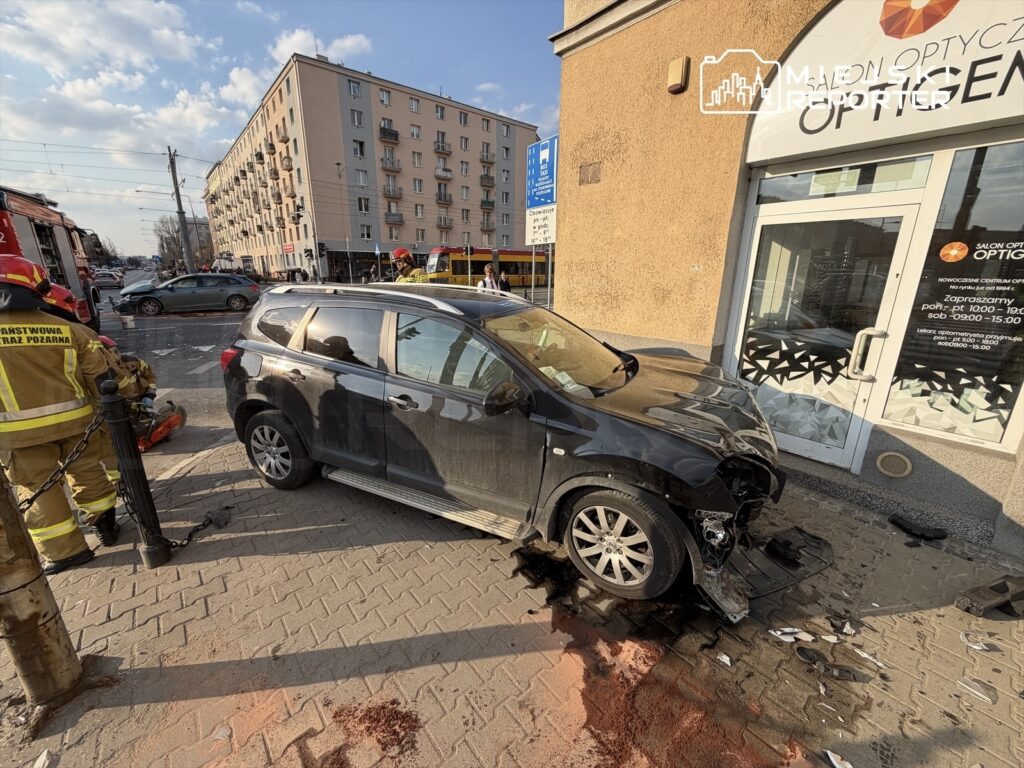 Czarny samochód osobowy z uszkodzoną przednią częścią stoi na chodniku, obok pracuje straż pożarna, w tle budynki mieszkalne.