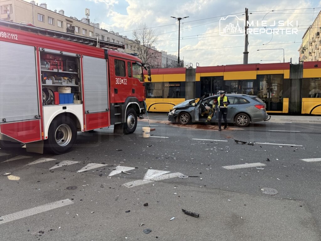Strażak przy wozie strażackim ocenia skutki wypadku drogowego obok uszkodzonego samochodu i tramwaju.