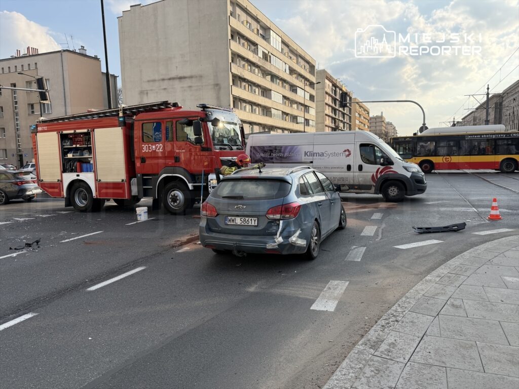 W miejscu wypadku stoi wóz strażacki, obok uszkodzony samochód osobowy, na drodze widoczne są resztki pojazdów.