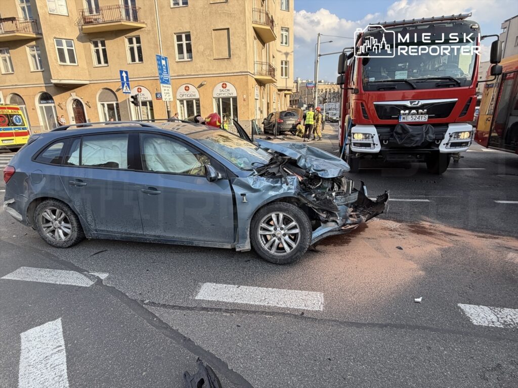 Rozbity samochód osobowy z uszkodzoną przednią częścią na skrzyżowaniu, obok strażackiego wozu ratunkowego.