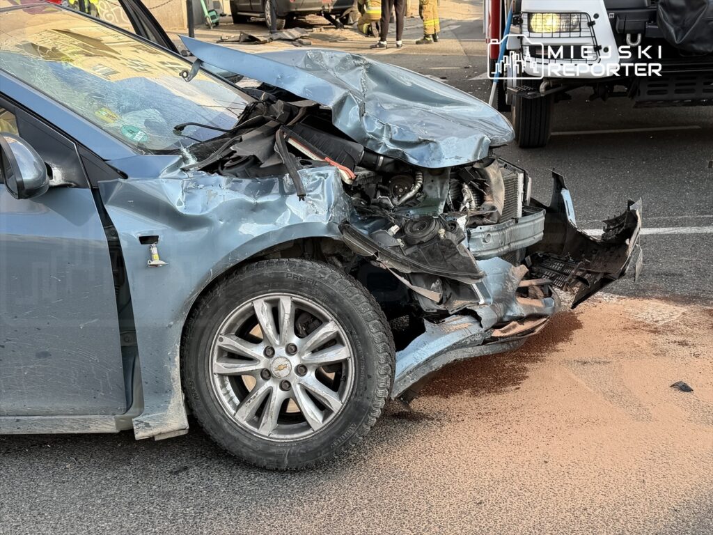 Uszkodzony samochód osobowy z wgniecionym przodem leży na jezdni, obok rozlane substancje płynne.