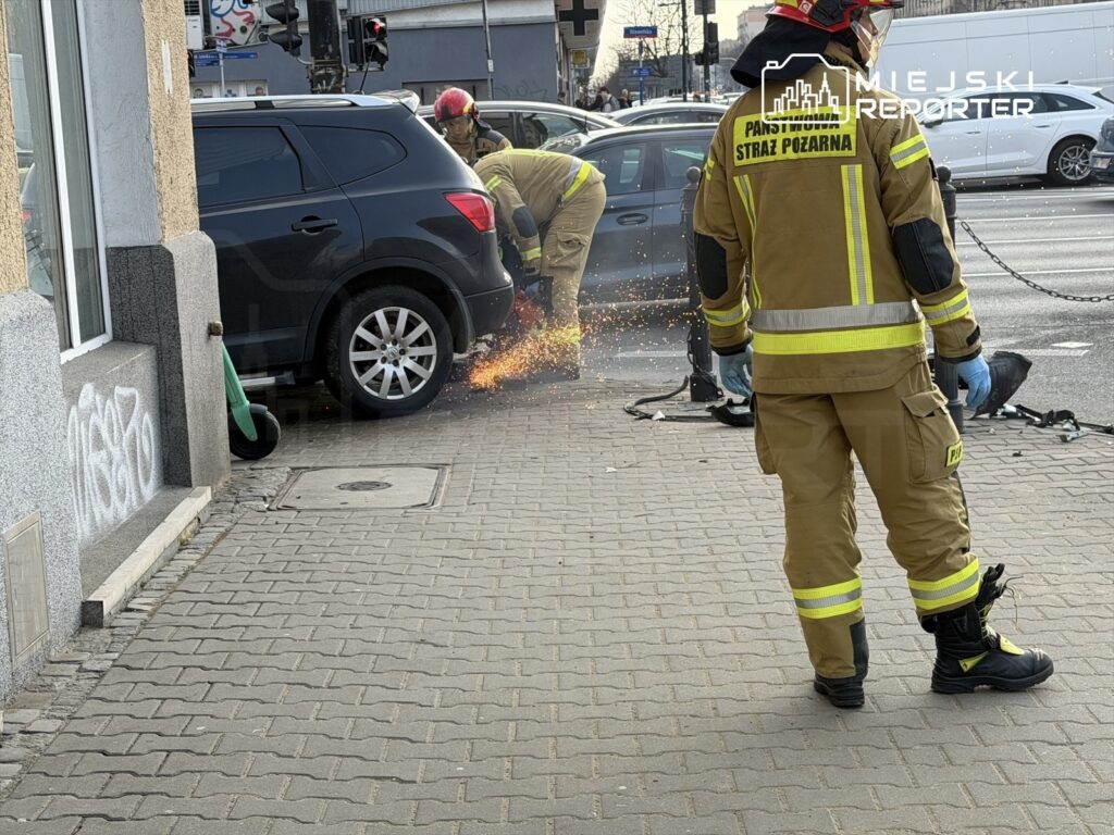 Strażacy w mundurach tną metal w pobliżu zaparkowanego SUV-a, generując iskry na chodniku.