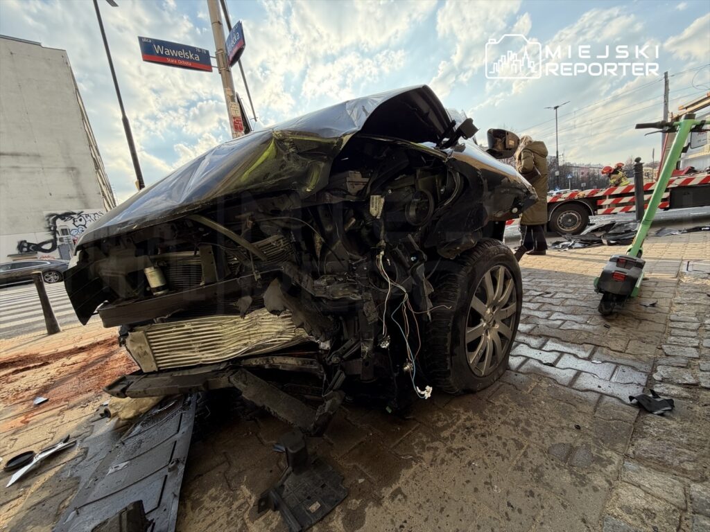 Zniszczony samochód osobowy leży na chodniku, obok widać uszkodzenia oraz pracowników służb ratunkowych.