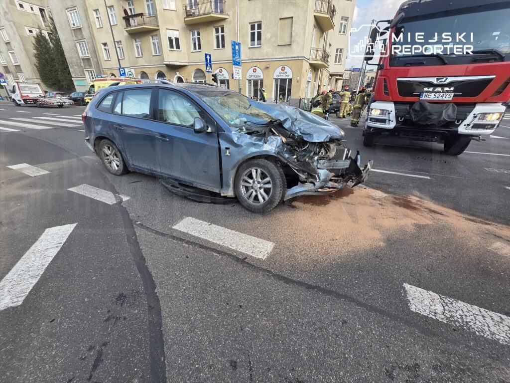 Zniszczony samochód osobowy leży na jezdni po wypadku, obok wozu strażackiego i grupy ratowników.