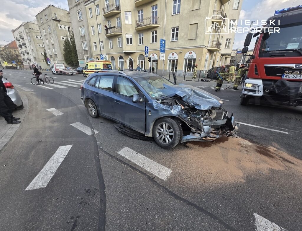 Uszkodzony samochód osobowy z wgniecionym przodem stoi na jezdni, obok interweniują strażacy i karetka.