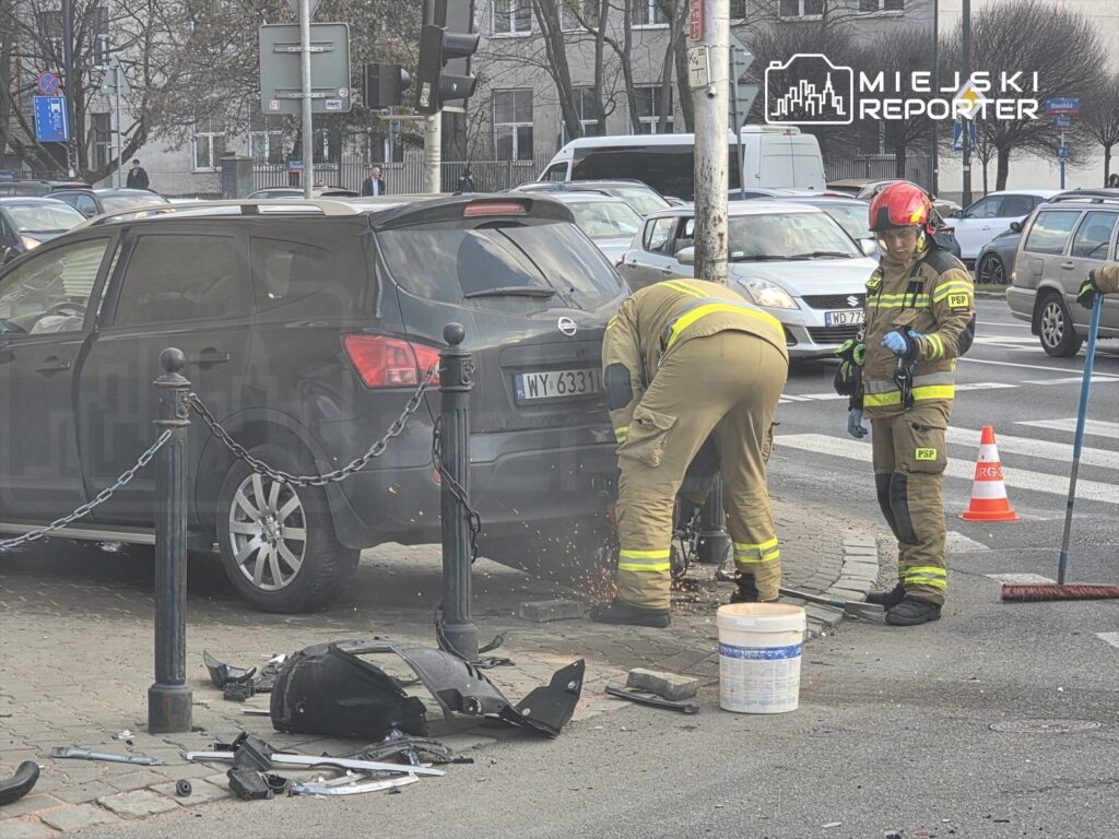 Strażacy usuwają zniszczoną część samochodu na ulicy, obok widać porozrzucane elementy pojazdu.