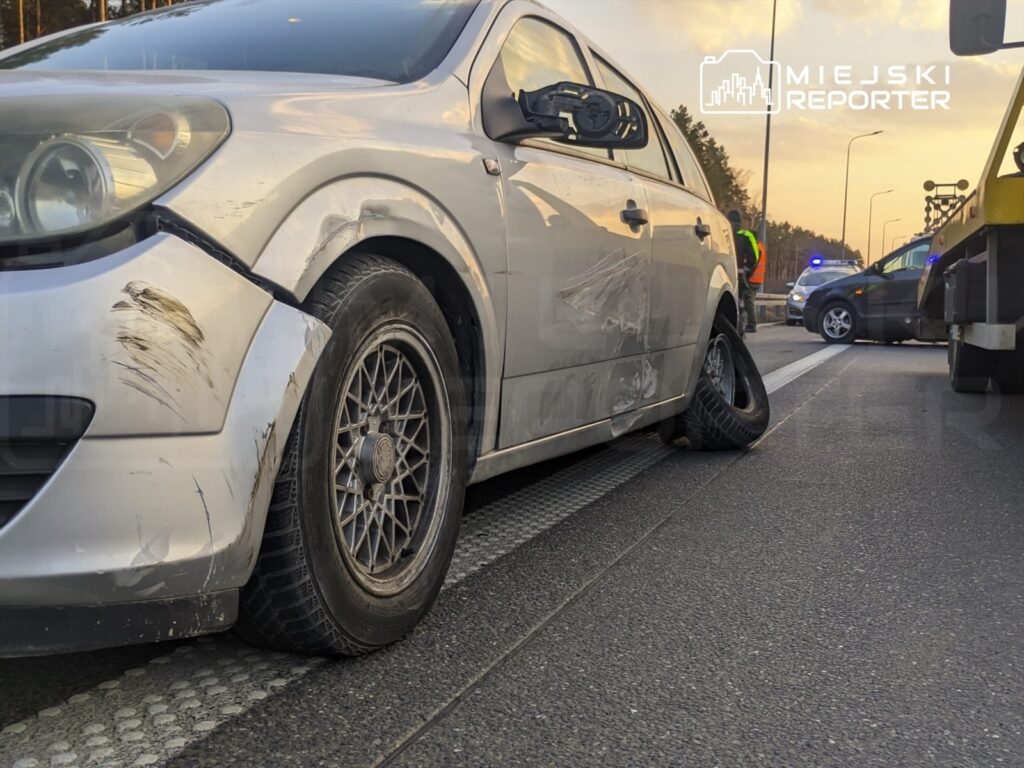 Uszkodzony samochód osobowy z zarysowaniami na karoserii i przebitą oponą na drodze, obok stoi laweta.