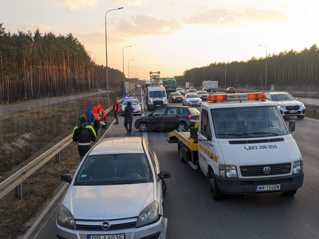 Grupa osób w kamizelkach odblaskowych udziela pomocy na poboczu drogi, obok stoi laweta z napisem "POMOC DROGOWA".