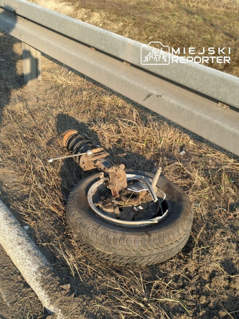 Zniszczone koło z elementami zawieszenia leżące na poboczu drogi, obok bariery ochronnej.