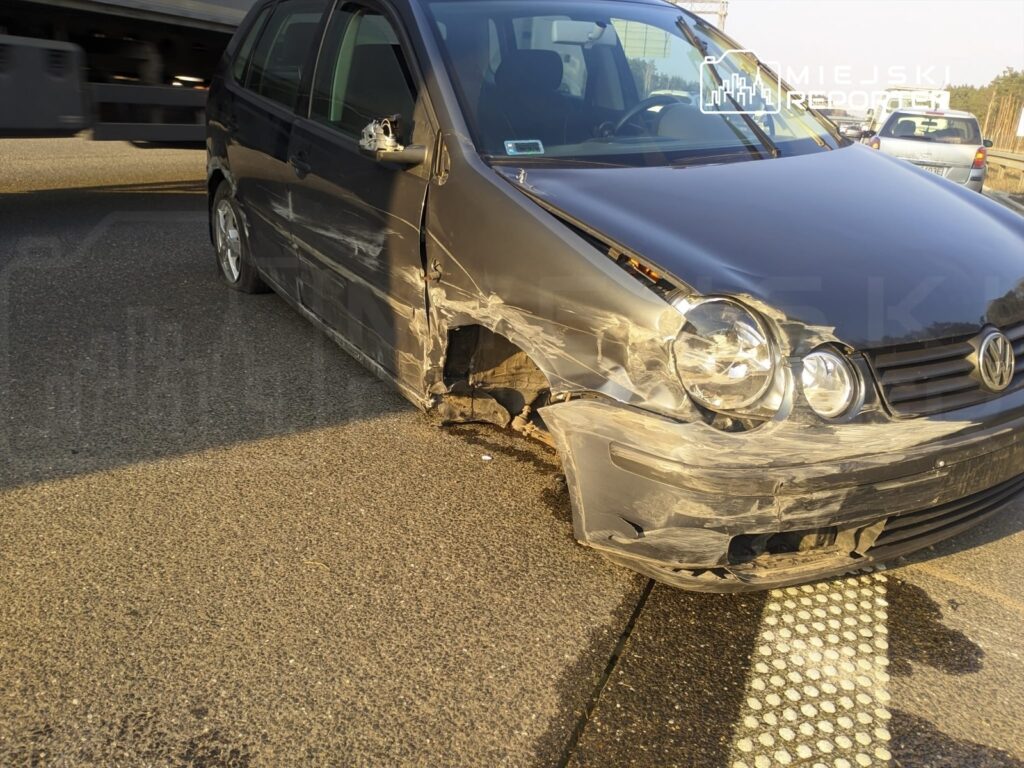 Uszkodzony samochód osobowy zaparkowany na drodze, z widocznymi zniszczeniami z lewej strony.