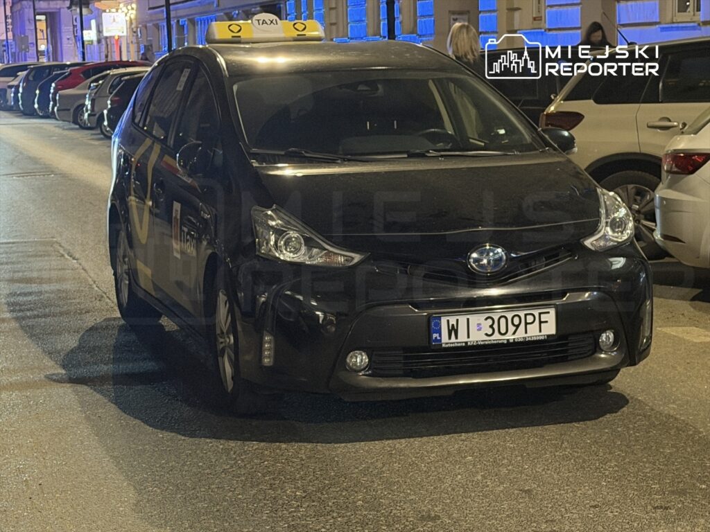 Czarny taksówka zaparkowana na oświetlonej ulicy, obok innych samochodów, z widocznym znakiem "TAXI" na dachu.